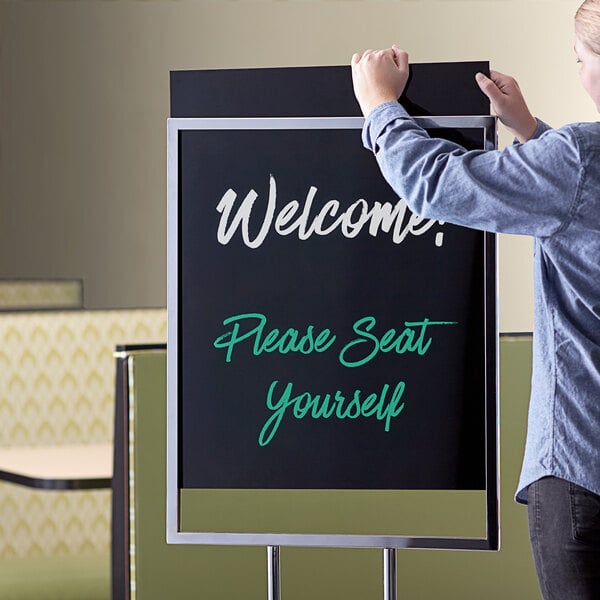 A woman standing in front of a black double sided Aarco marker board with white writing on it.