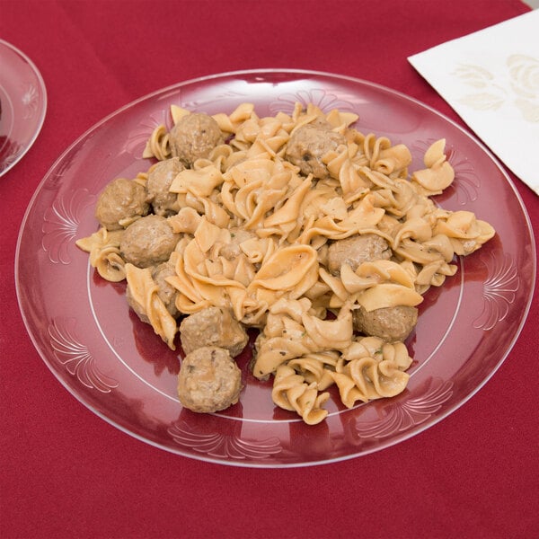 A WNA Comet clear plastic plate with a white design holding pasta and meatballs on a red tablecloth.