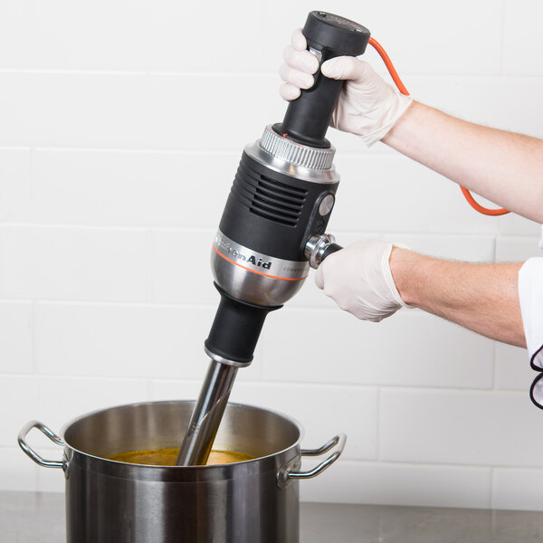A person using a KitchenAid 400 Series immersion blender to mix soup in a metal container.