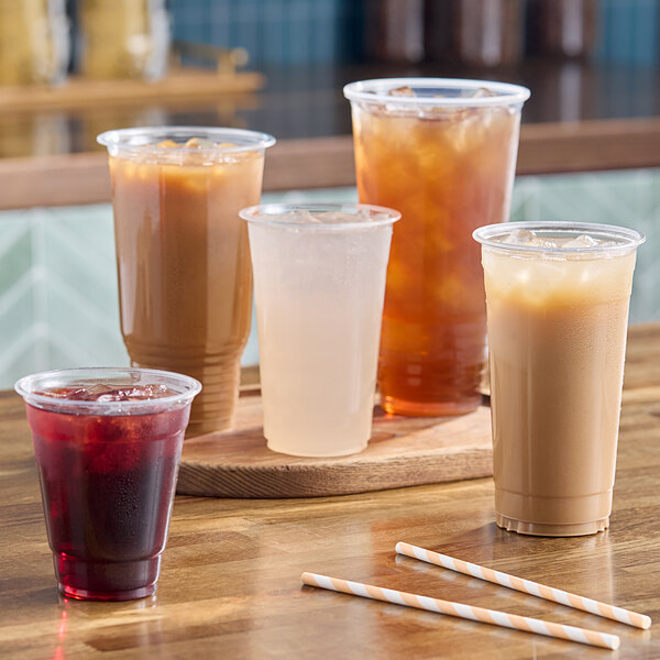 Five clear plastic cups filled with various iced beverages are arranged on a wooden surface, with three striped paper straws placed in front.