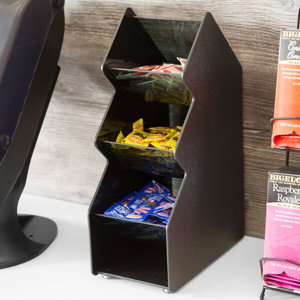 A black metal Vollrath 3-bin condiment holder on a shelf with candy in it.