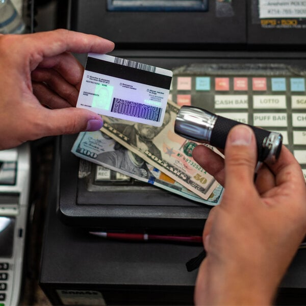 A person is using a handheld UV light to check the authenticity of an identification card near a cash register with cash visible underneath.