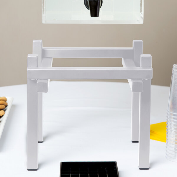 A white table with a white metal Cal-Mil beverage dispenser base holding a glass of water.