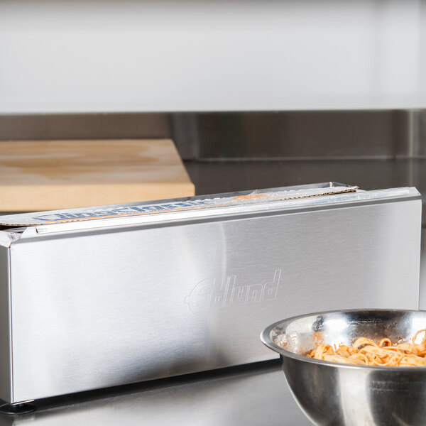 A silver metal Edlund foil dispenser blade in a metal box on a counter with a bowl of pasta.