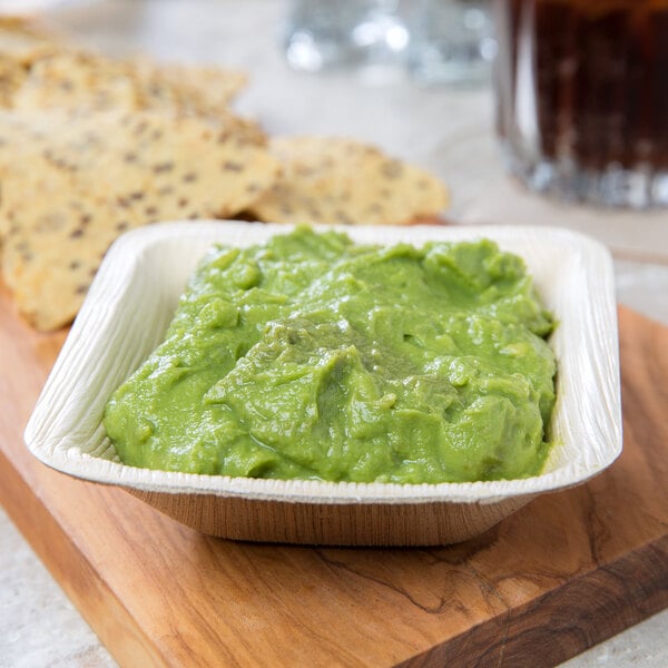 A 5 oz. square palm leaf bowl filled with guacamole on a cutting board.