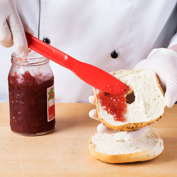 A person using an HS Inc. red polypropylene sandwich spreader to spread jam on a bagel.