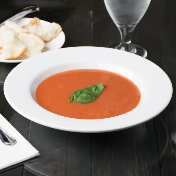 A Libbey aluma white porcelain deep soup bowl with soup next to a plate of bread and a spoon.