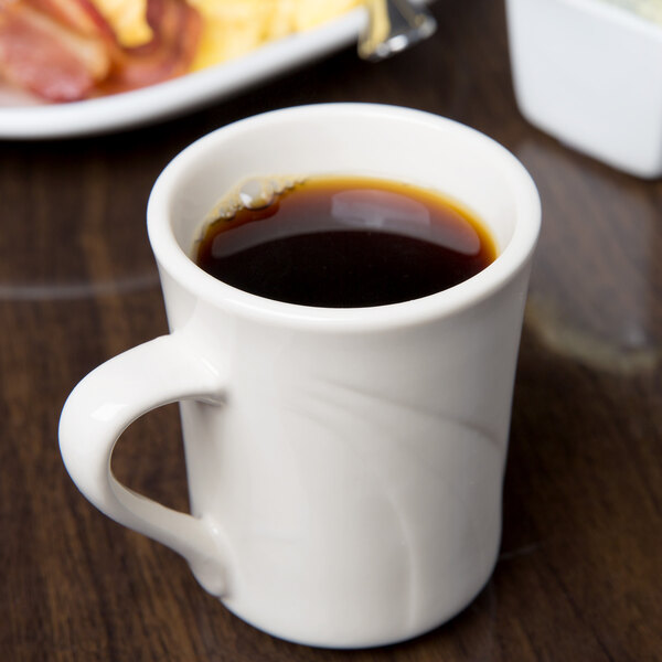 A Libbey ivory porcelain mug filled with brown liquid on a table with a plate of bacon.