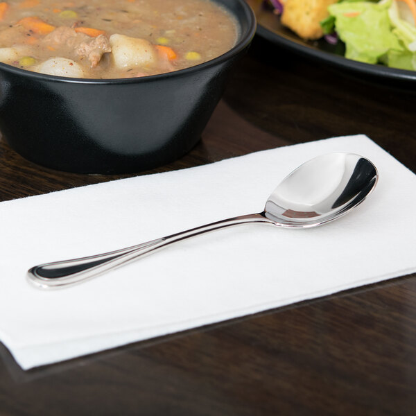 A Libbey stainless steel bouillon spoon on a napkin next to a bowl of soup.