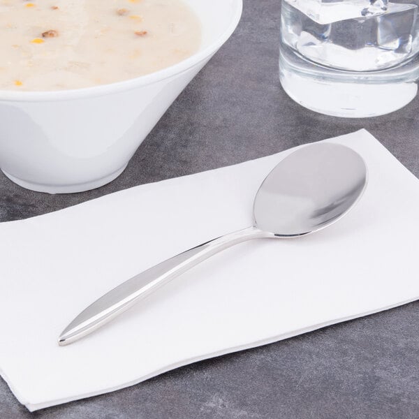 A Libbey stainless steel bouillon spoon on a napkin next to a bowl of soup.