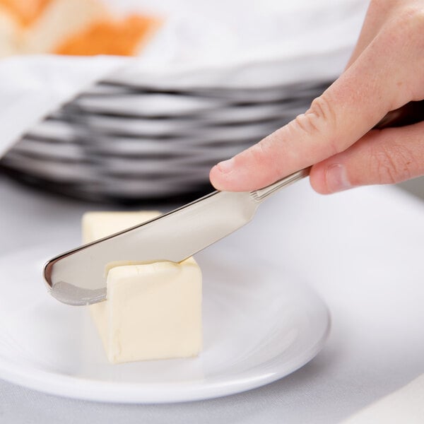 A person's hand holding a Libbey Geneva stainless steel butter spreader over a piece of butter on a plate.