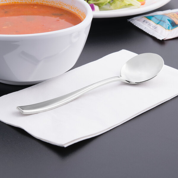 A Libbey stainless steel bouillon spoon on a napkin next to a bowl of soup.