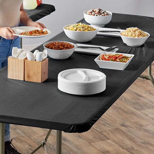 A woman using a black rectangular plastic tablecloth with elastic to serve food on a table.