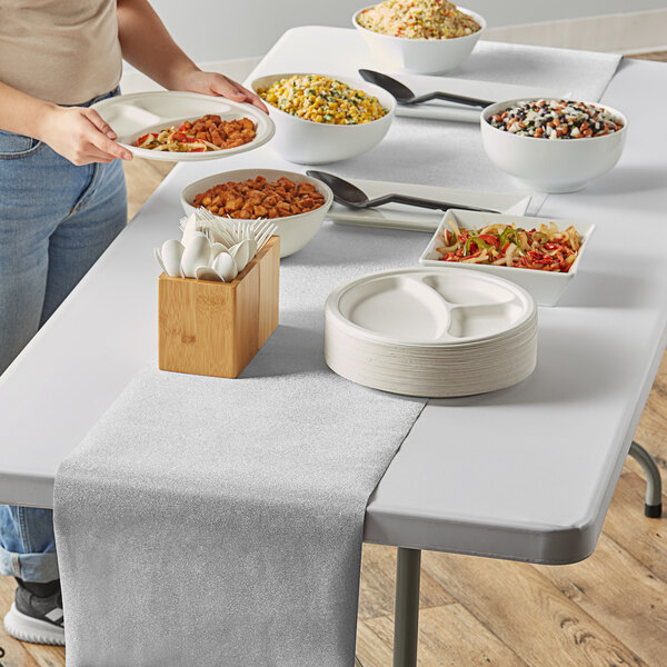 A woman serving food on a table with a Creative Converting silver glitter plastic table runner next to a bowl of food and stack of plates.