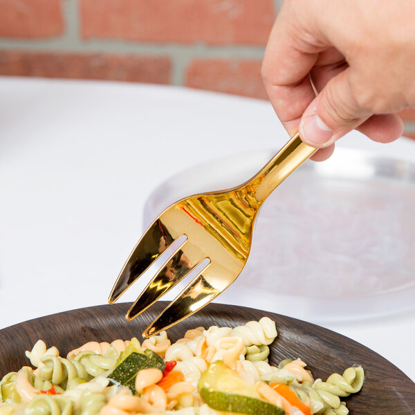 A hand holding a Fineline Golden Secrets gold serving fork over a plate of pasta.