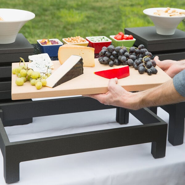 A hand holding a Vollrath cutting board with grapes and cheese on a table.