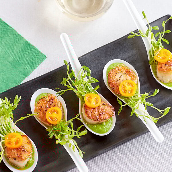 A Visions white plastic Asian soup spoon full of scallops on a plate with tomato and greens.