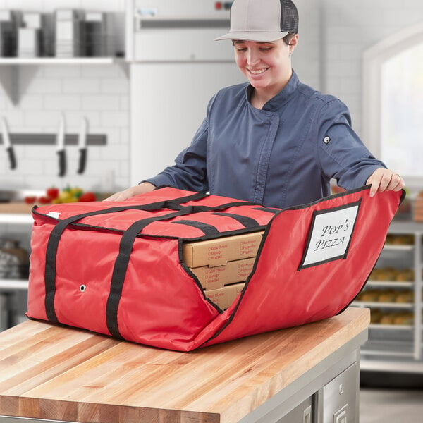 A woman in a blue jacket and chef hat holding a red CaterGator Dash insulated pizza delivery bag.