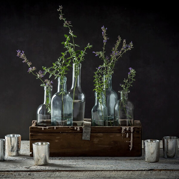 A clear glass olive oil cruet with a pourer, displayed among other glass bottles and greenery in a rustic wooden box.