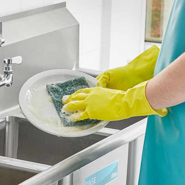 A person wearing yellow Cordova Latex rubber gloves washing a dish with a sponge.