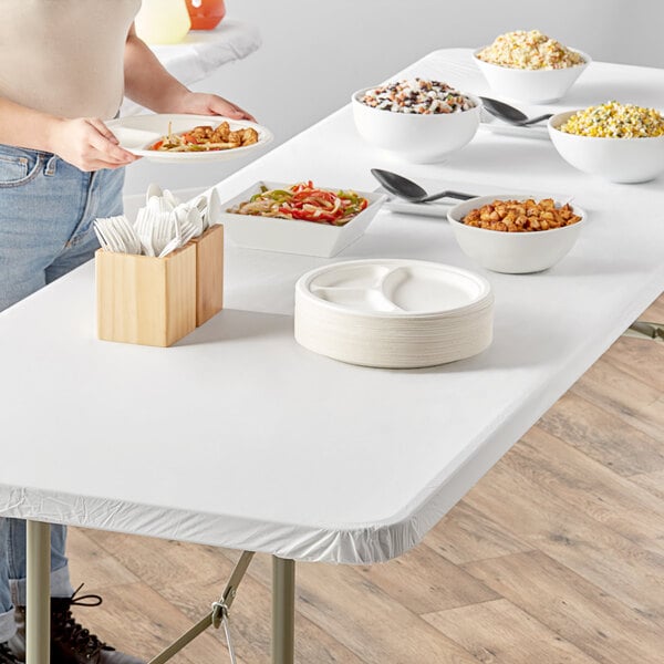 A woman standing at an outdoor table with white Creative Converting Stay Put tablecloth filled with food including rice and vegetables.