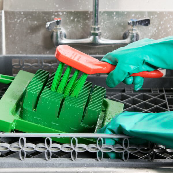 A person in green gloves uses a Prince Castle Saber King cleaning brush to clean a dishwasher.