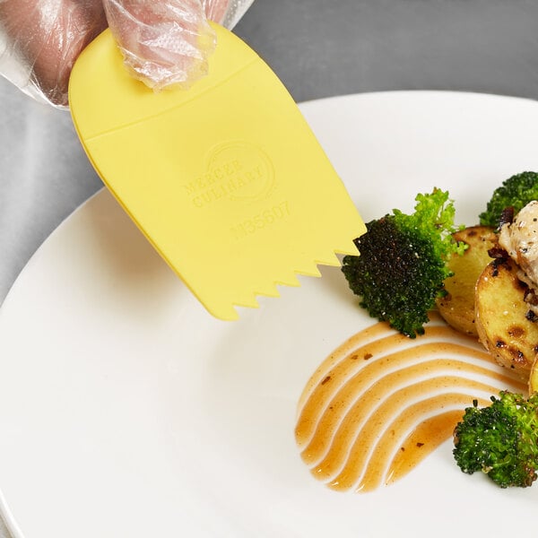 A person using a yellow Mercer Culinary silicone wedge to plate green and black vegetables.