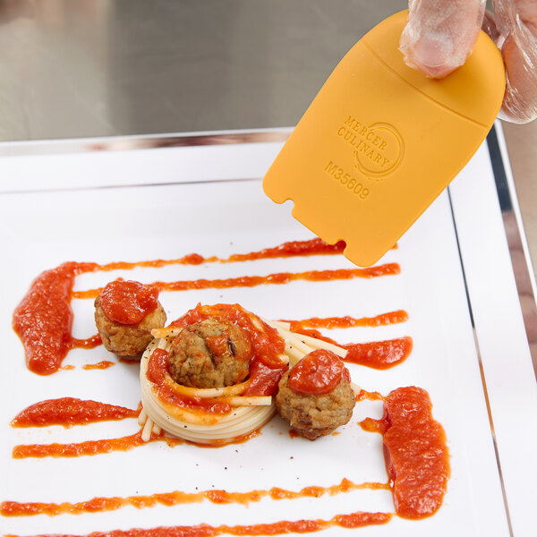 A hand using a Mercer Culinary horseshoe arch silicone wedge plating tool to add sauce to a plate of food.