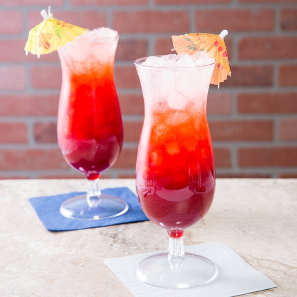 Two Fineline Quenchers plastic hurricane glasses filled with red and yellow drinks with umbrellas on a table.