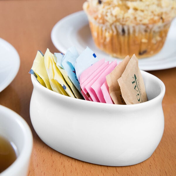 A Libbey Royal Rideau white porcelain sugar packet holder on a table with a bowl of sugar packets.