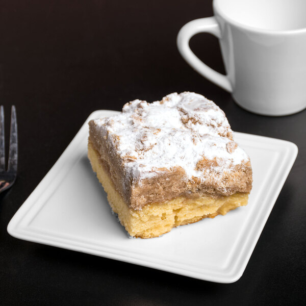 A piece of cake on a Libbey white porcelain square plate next to a fork and a white mug.