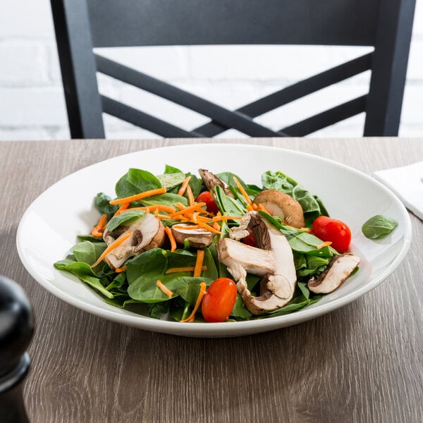A white round porcelain bowl filled with a fresh salad containing spinach, mushrooms, cherry tomatoes, and shredded carrots.