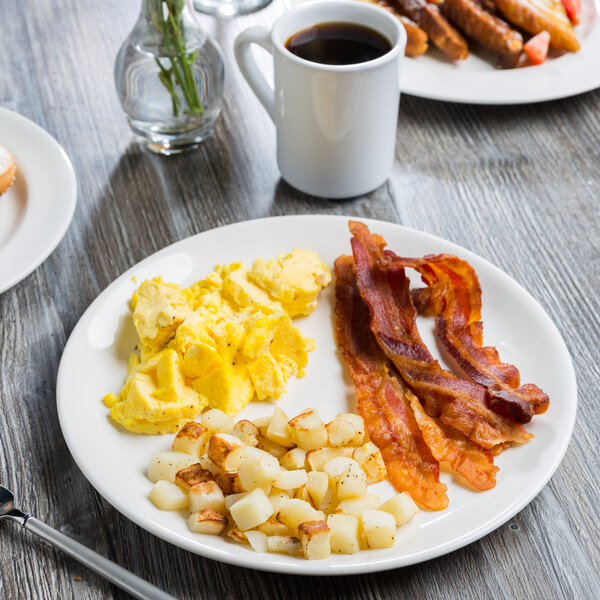 A Libbey Royal Rideau white porcelain coupe plate with eggs, bacon, and potatoes on a table.