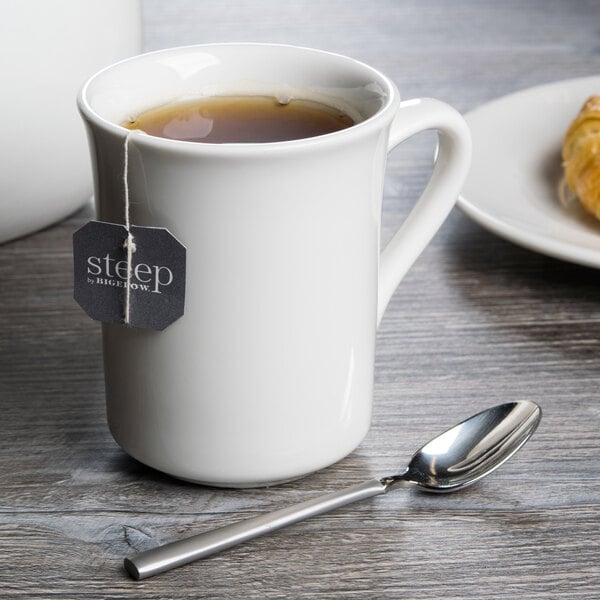 A white Libbey porcelain mug filled with tea and a tea bag with a spoon next to it.