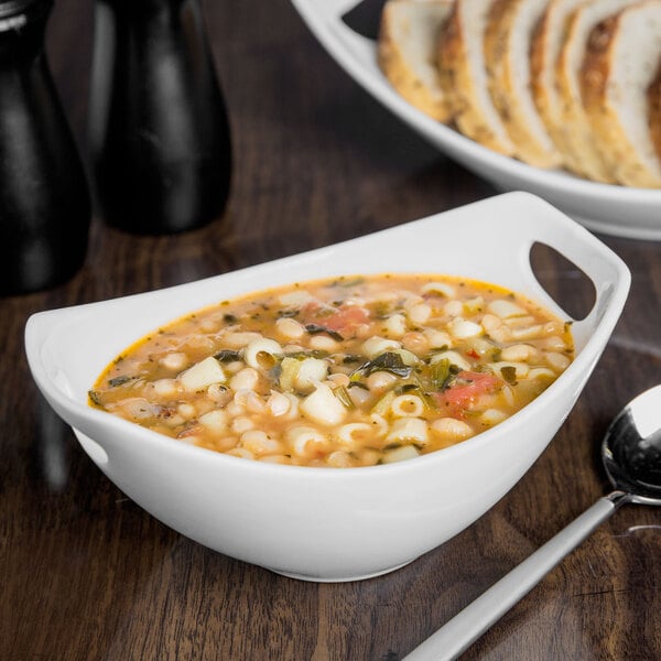 A Libbey white porcelain bowl filled with soup and pasta on a table with bread.