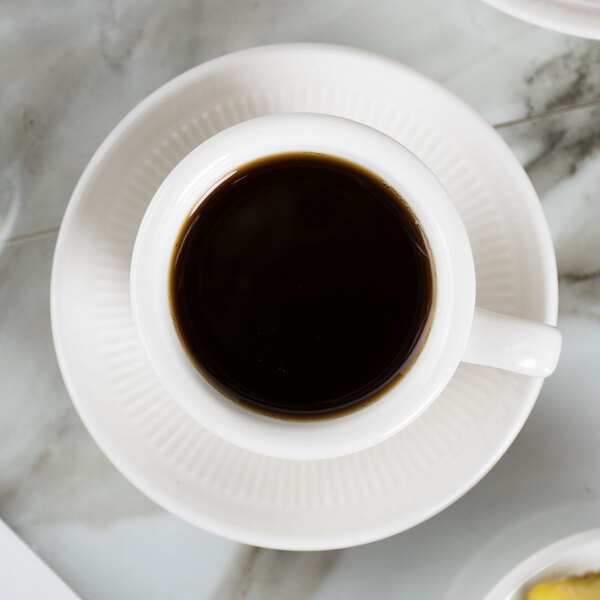 A cup of coffee sits on a Tuxton Hampshire saucer on a table.