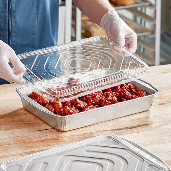 A person in gloves holding a Durable Packaging aluminum tray of food with a clear plastic dome lid.