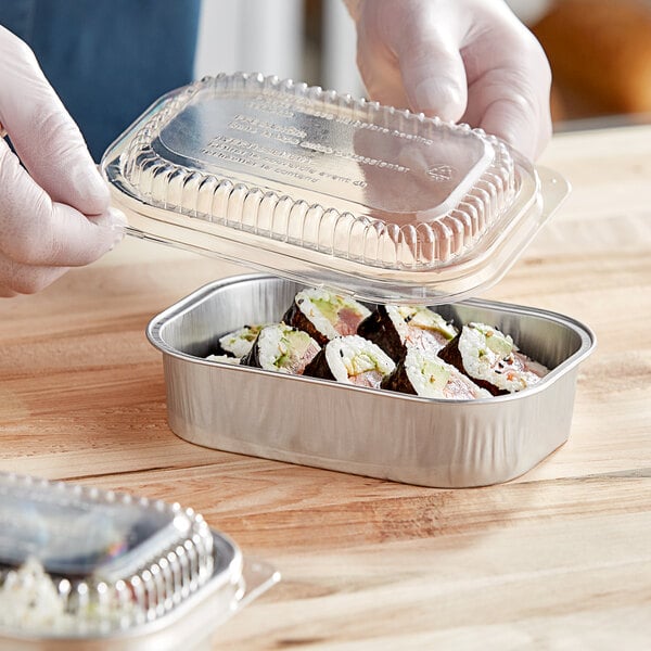 A hand holding a clear plastic container of sushi with a silver lid.