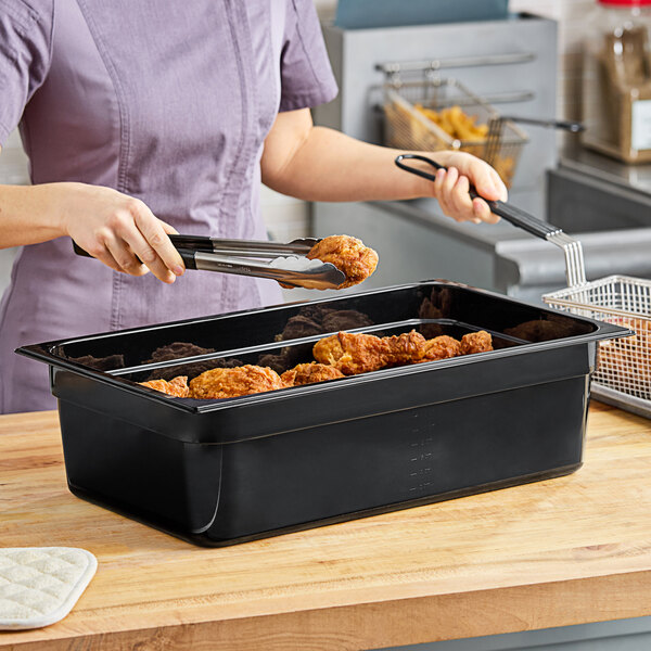 A woman holding tongs over a black Carlisle plastic food pan filled with fried chicken.