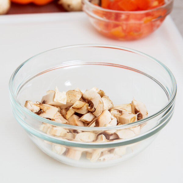 An Arcoroc glass bowl of sliced mushrooms on a cutting board.