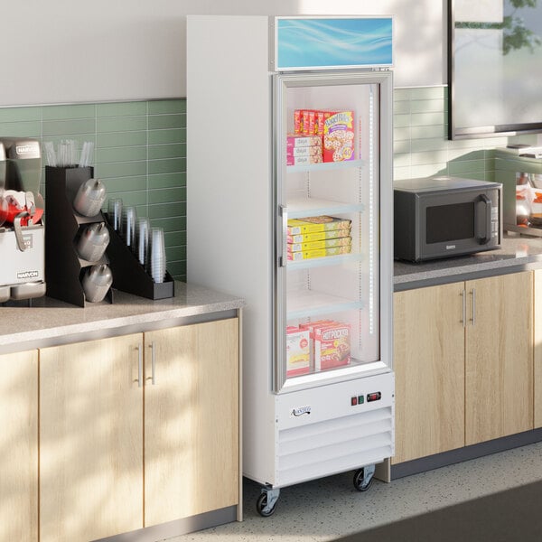 A white glass door merchandiser freezer with LED lighting, stocked with frozen food items and positioned between countertops in a commercial kitchen or break room.
