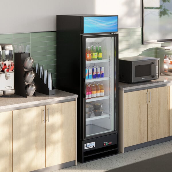 A black glass door merchandiser refrigerator with LED lighting, stocked with beverages and food items, situated between a microwave and a drink station in a break room setting.