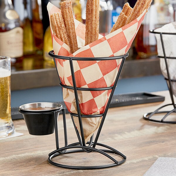 A Clipper Mill black wire cone basket with a churro and a cup on a table with a red and white checkered bag inside.