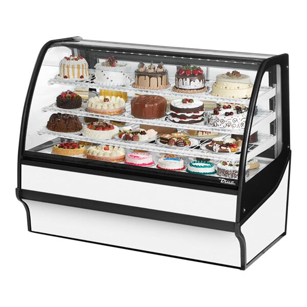 A True white curved glass refrigerated bakery display case full of cakes with frosting and fruit decorations.