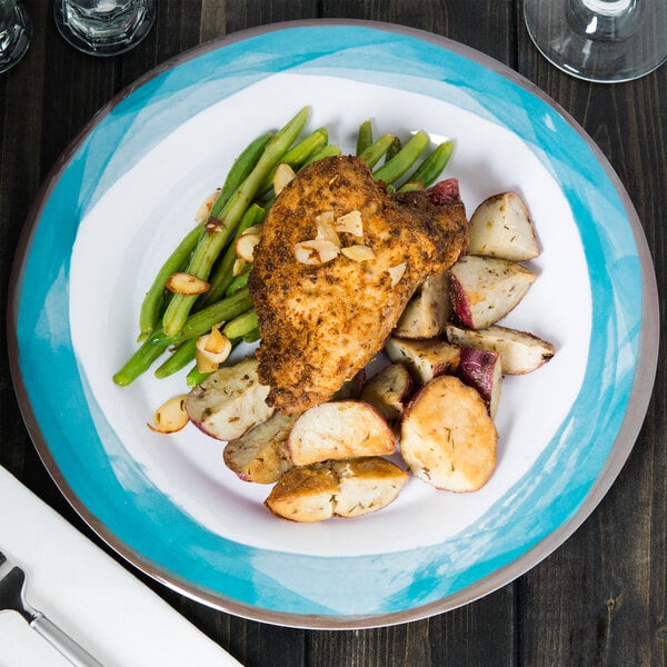 A Kanello white melamine plate with a Kanello blue edge on a table with a plate of chicken, potatoes, and green beans with a fork and knife.