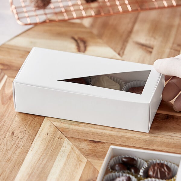 A hand holding a 7 1/8" x 3 3/8" white candy box with a triangular window containing chocolates.