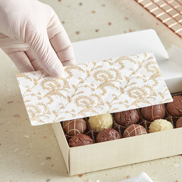 A hand in a glove taking a gold and white floral patterned pad out of a white candy box.