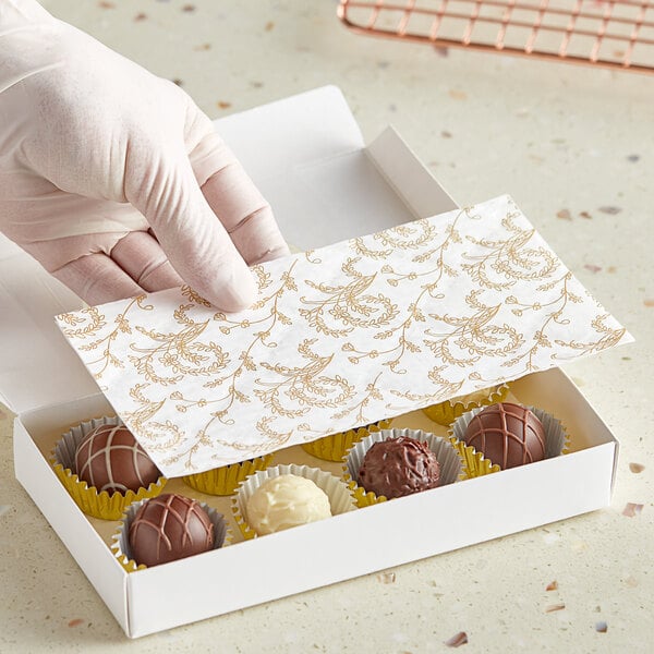 A hand in a white glove holding a glassine paper over a white candy box with a gold floral pattern.