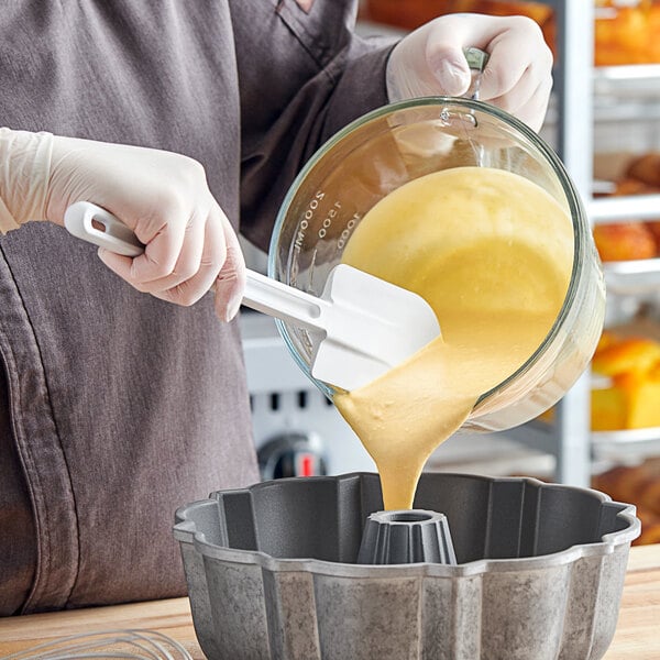 A hand using a Rubbermaid white spatula to pour batter into a cake mold.