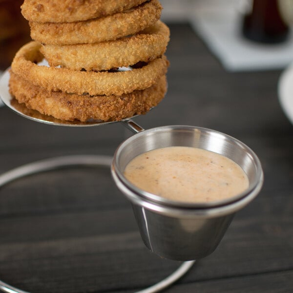 A Clipper Mill stainless steel onion ring tower with two ramekin holders holding a stack of fried onion rings on a metal tray with sauce.